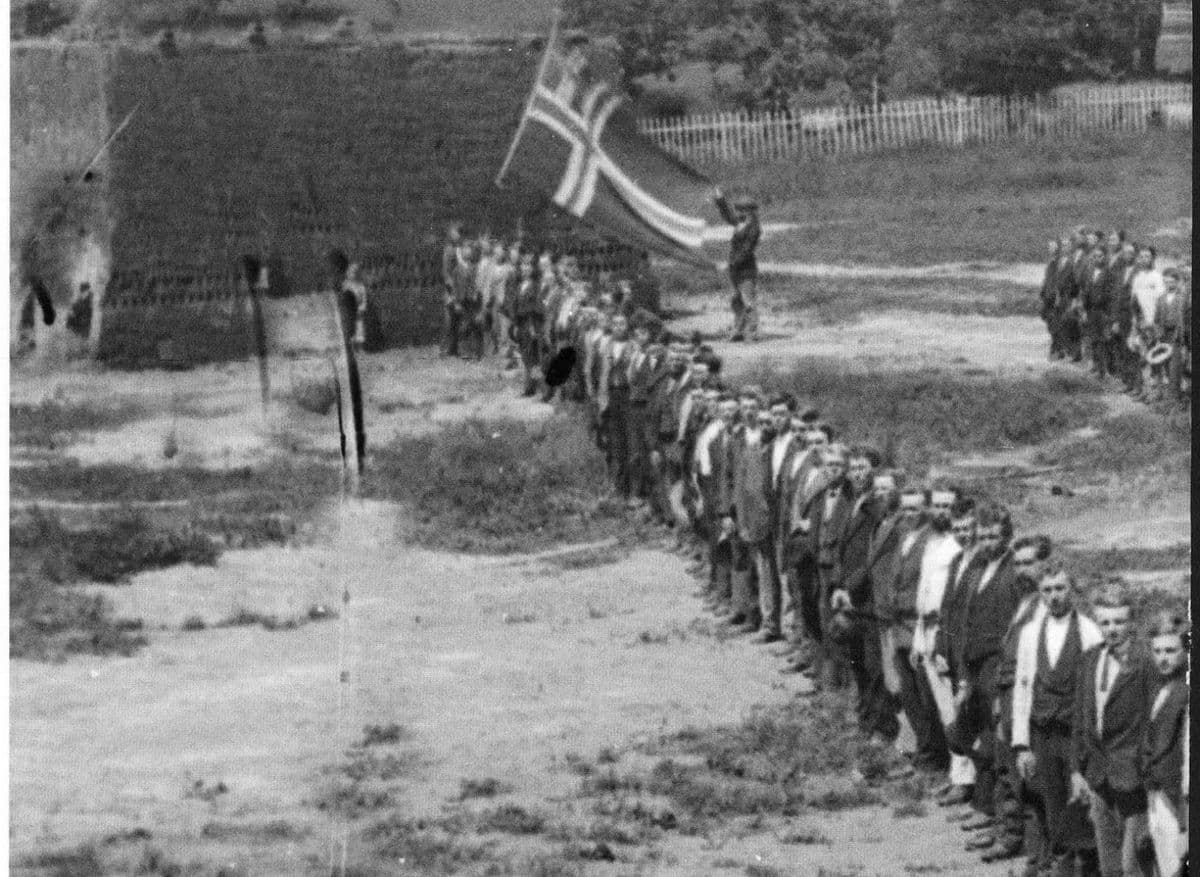 Oppstilte frivillige under norsk flagg ved en murbygning, historisk svart-hvitt-foto